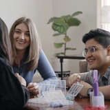 Foster mom and dad sitting at table making beaded bracelets with young foster daughter.